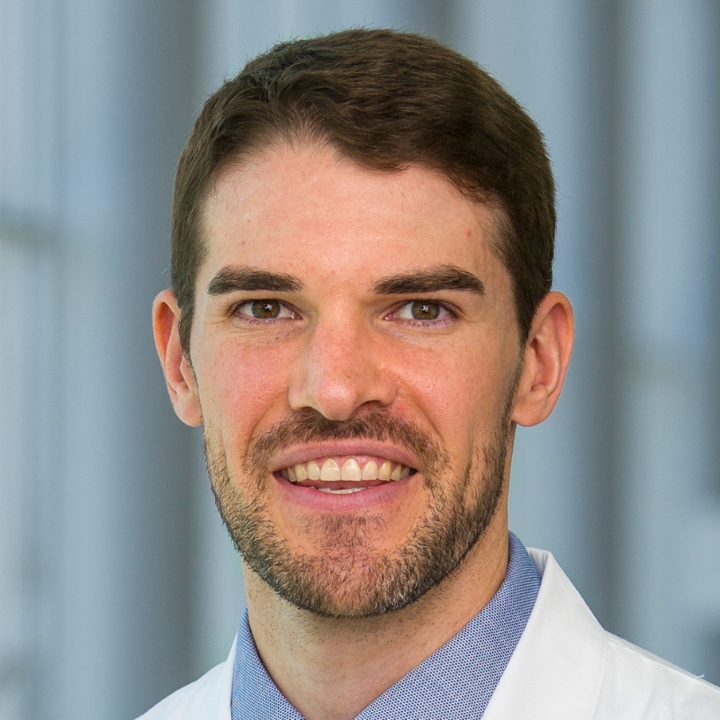 Smiling man with brown hair, mustache, and beard wearing a light blue dress shirt, blue and red tie, and a white lab coat